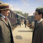 Zwei Männer in Anzügen stehen an einem Bahnsteig, es gibt ein großes Schild mit der Aufschrift "RIVA" im Hintergrund.