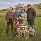 Eine Gruppe von fünf Personen steht auf einer Wiese neben einem Holzhandwagen, die Kleidung wirkt ländlich und von früher.