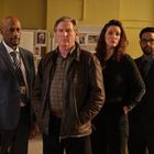 Vier ernst dreinblickende Menschen in einem Büro, mit einer Tafel im Hintergrund und Computern.