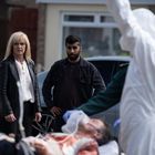 Zwei Menschen schauen ernst auf einen Mann auf einer Trage, während ein Helfer in Schutzkleidung daneben steht.