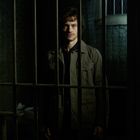 Keine sichtbaren Texte. Ein Mann steht hinter Gittern in einem düsteren Raum mit Toilette und Ziegelwand.