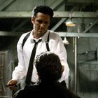 Ein Mann im weißen Hemd mit Krawatte und Hosenträgern hält ein Messer in einer düsteren Halle vor einem sitzenden Mann.