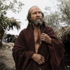 Ein Mann mit Bart und langen Haaren trägt einen braunen Umhang. Im Hintergrund sind rustikale Hütten und Bäume erkennbar.