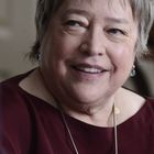 Eine ältere Frau mit kurzem grauem Haar lächelt freundlich. Sie trägt eine rote Bluse und Ohrringe.