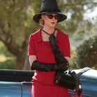 Eine Frau in einem roten Outfit, schwarzer Hut und Handschuhe, steht neben einem blauen Auto. Sie trägt eine Sonnenbrille.