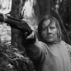 Eine Person in einem Wald zielt mit einer Flinte. Sie trägt robuste Kleidung und wirkt entschlossen.