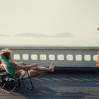 Eine Frau im roten Kleid steht auf einem Schiffdeck; zwei Personen entspannen auf Sonnenliegen. Inseln im Hintergrund.