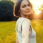 Die Szene zeigt eine Frau auf einer Wiese bei Sonnenuntergang. Sie blickt über die Schulter, hält eine Hand, Bäume im Hintergrund.