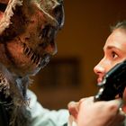 Kein Text erkennbar. Eine Person mit gruseliger Maske hält eine zweite Person, die eine Pistole in der Hand hat.