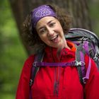 Eine Person mit roter Jacke und lila Stirnband lächelt im Wald. Sie trägt einen Wanderrucksack.