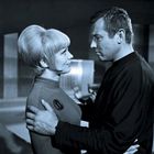 Eine Frau und ein Mann stehen sich in einem futuristischen Raum nah gegenüber. Die Frau trägt ein Emblem.