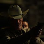 Ein Mann in einem Cowboyhut zielt mit einer Pistole, im Hintergrund ist es dunkel.