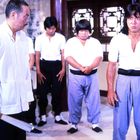 Fünf Männer in Kampfkunstkleidung, ernst schauend. Einer hält einen Fächer, Szene in traditionell dekoriertem Raum.