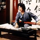 Chinesische Schriftzeichen auf einer Wand. Ein Mann in grauer Kleidung sitzt, hält einen roten Schirm und sieht entschlossen aus.