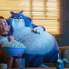 Ein ängstlicher Mann sitzt mit einer riesigen, fröhlichen Katze auf einem Sofa. Beide halten eine Schüssel Popcorn.