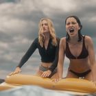 Zwei Frauen auf gelber Luftmatratze im Wasser. Eine mit schwarzem Oberteil, die andere in einem braunen Bikini. Dramatischer Himmel.