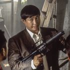 Ein Mann in einem Anzug hält ein Gewehr mit Schalldämpfer, spricht mit zwei anderen, Szene in einem Lagerraum.