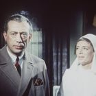 Ein Mann im Anzug mit Krawatte steht neben einer lächelnden Frau in einer Krankenschwesteruniform. Im Hintergrund ein Bild.