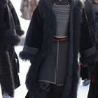 Eine Person im langen, schwarzen Mantel mit Fell. Dahinter verschneite Landschaft und unscharfe Figuren in Winterkleidung.