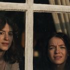 Zwei Frauen schauen durch ein viergeteiltes Fenster. Eine wirkt nachdenklich, die andere besorgt. Im Hintergrund sind Vorhänge.
