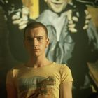 Vorne steht ein Mann mit gelbem T-Shirt, darauf ein Strandmotiv. Dahinter ein Gemälde mit einer Person und zwei Waffen.