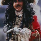 Ein Mann in einem aufwendigen Piratenkostüm mit rotem Mantel und Hakenhand lächelt. Er trägt einen großen verzierten Hut.