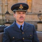 Ein Mann in einer blauen Uniform mit Mütze, sitzt vor einem alten Gebäude. Im Hintergrund ein Militärfahrzeug.