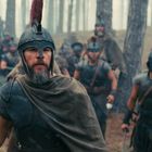 Ein Krieger mit Helm und rotem Kamm führt eine Gruppe bewaffneter Männer in einem nebligen Wald an.
