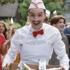 Ein fröhlicher Mann in einer weißen Uniform mit roter Fliege verteilt Getränke an lachende Kinder im Hintergrund.