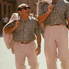Zwei lachende Männer in identischen Outfits mit Sonnenbrillen, einer groß, der andere klein, gehen nebeneinander.