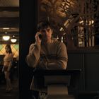 Ein Mann in einem gestreiften Pullover telefoniert, stehend vor dekorativer Wand. Im Hintergrund ein Restaurant mit Gästen.