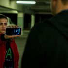 Text "ELEVATOR" auf gelbem Schild, Frau in roter Jacke zeigt Handy in Tiefgarage, Mann von hinten zu sehen.