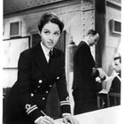 Eine Frau in Uniform beugt sich über einen Tisch und schreibt. Im Hintergrund stehen zwei Männer in Uniform.