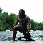 Samson kniet in einem flachen Fluss, umgeben von Wald, wild und kraftvoll aussehend, mit zerzausten Haaren und Narben.