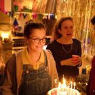 "ANNIVERSARY" auf einem Banner. Eine lachende Frau hält eine Geburtstagstorte mit brennenden Kerzen, umgeben von anderen Leuten.