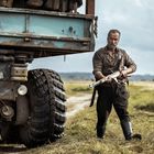 Ein Mann mit Gewehr unter einem großen, alten Lastwagen auf einem Feldweg, der Himmel ist bewölkt.