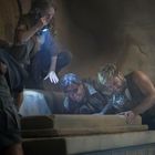 Vier Personen heben einen schweren Steinblock in einer höhlenartigen Umgebung an. Eine Frau hält eine Taschenlampe.