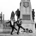 Zwei Männer tanzen humorvoll vor einem Kriegsdenkmal mit Soldatenstatuen und Kränzen. Hintergrund: Gebäude und Statue.