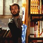 Ein Mann mit Bart schaut neugierig zwischen Bücherregalen hervor. Im Hintergrund sind Bücher mit bunten Buchrücken.