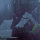 Ein riesiger Dinosaurier, möglicherweise ein T-Rex, greift ein umgestürztes Fahrzeug im Regen an.