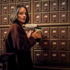 Eine Frau in einem langen Mantel öffnet eine Schublade eines alten Karteikastenmöbels.