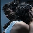 Zwei Personen umarmen sich emotional in einem schwach beleuchteten Raum. Bücher stehen im Hintergrund auf einem Regal.