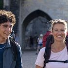 Ein lächelnder Mann und eine Frau mit Rucksäcken stehen vor einem steinernen Torbogen. Sie wirken fröhlich und entspannt.