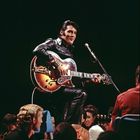 Ein Mann in schwarzer Lederjacke spielt Gitarre auf einer Bühne, umgeben von Publikum.