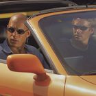 Zwei Personen mit Sonnenbrillen in einem orangefarbenen Sportwagen, die ernsthaft nach draußen schauen.