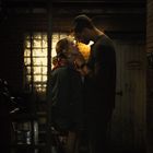 Text: "MOORE'S". Ein Paar steht nah beieinander in einem schmalen Ziegelkorridor, beleuchtet von schwachem Licht.