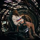 Person in Raumanzug sitzt in einem Cockpit mit vielen leuchtenden Anzeigen und einem runden Fenster im Hintergrund.