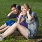 Zwei Frauen machen Sit-ups im Park. Eine trägt ein graues T-Shirt mit grünem Text, die andere ein lila Oberteil.