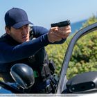 Ein Mann in dunkler Kleidung zielt mit einer Pistole, neben einem Auto, vor einer Landschaft mit grünem Gebüsch und blauem Himmel.