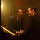 Zwei Männer in Anzügen sitzen an einem Klavier und lächeln sich in einem warm beleuchteten Raum an.